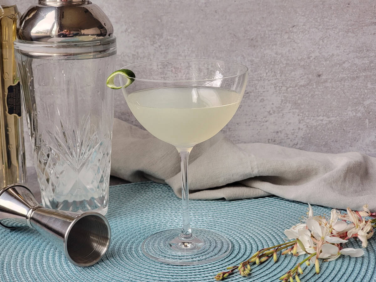 Hands shaking a metal cocktail shaker over a kitchen counter with limes and a jigger nearby