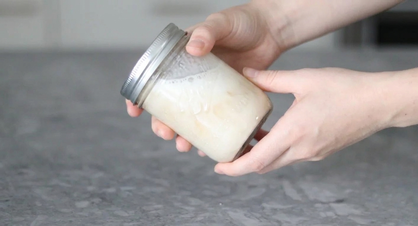 Hands shaking a sealed glass jar of creamy milk mixture over a kitchen counter