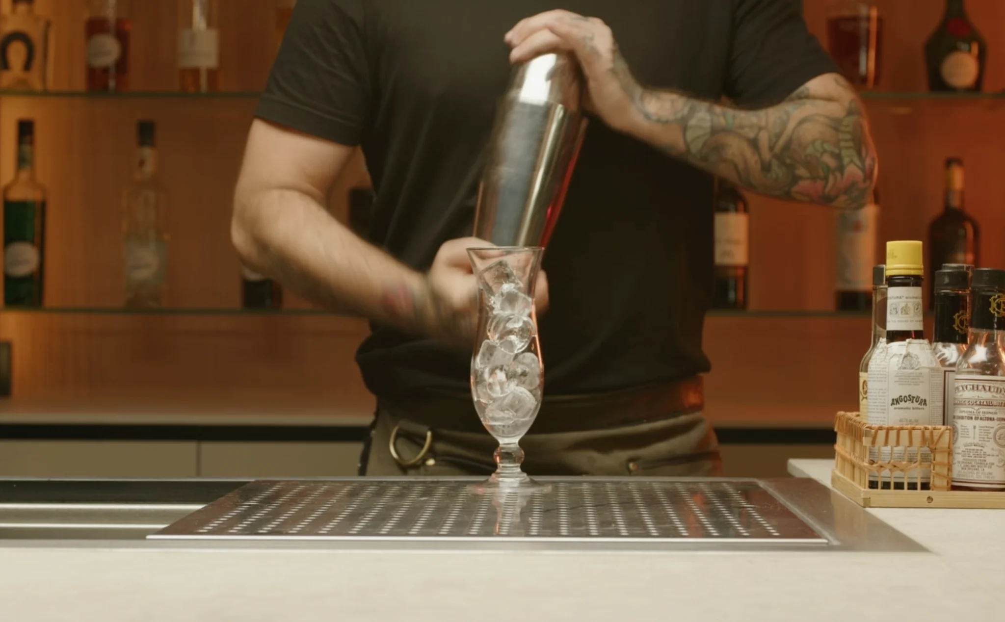 Hands shaking a stainless steel cocktail shaker over a kitchen counter with citrus halves nearby, mid-action in warm indoor lighting