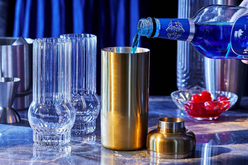 Hands shaking a stainless steel cocktail shaker over a light kitchen counter with a bowl of ice nearby