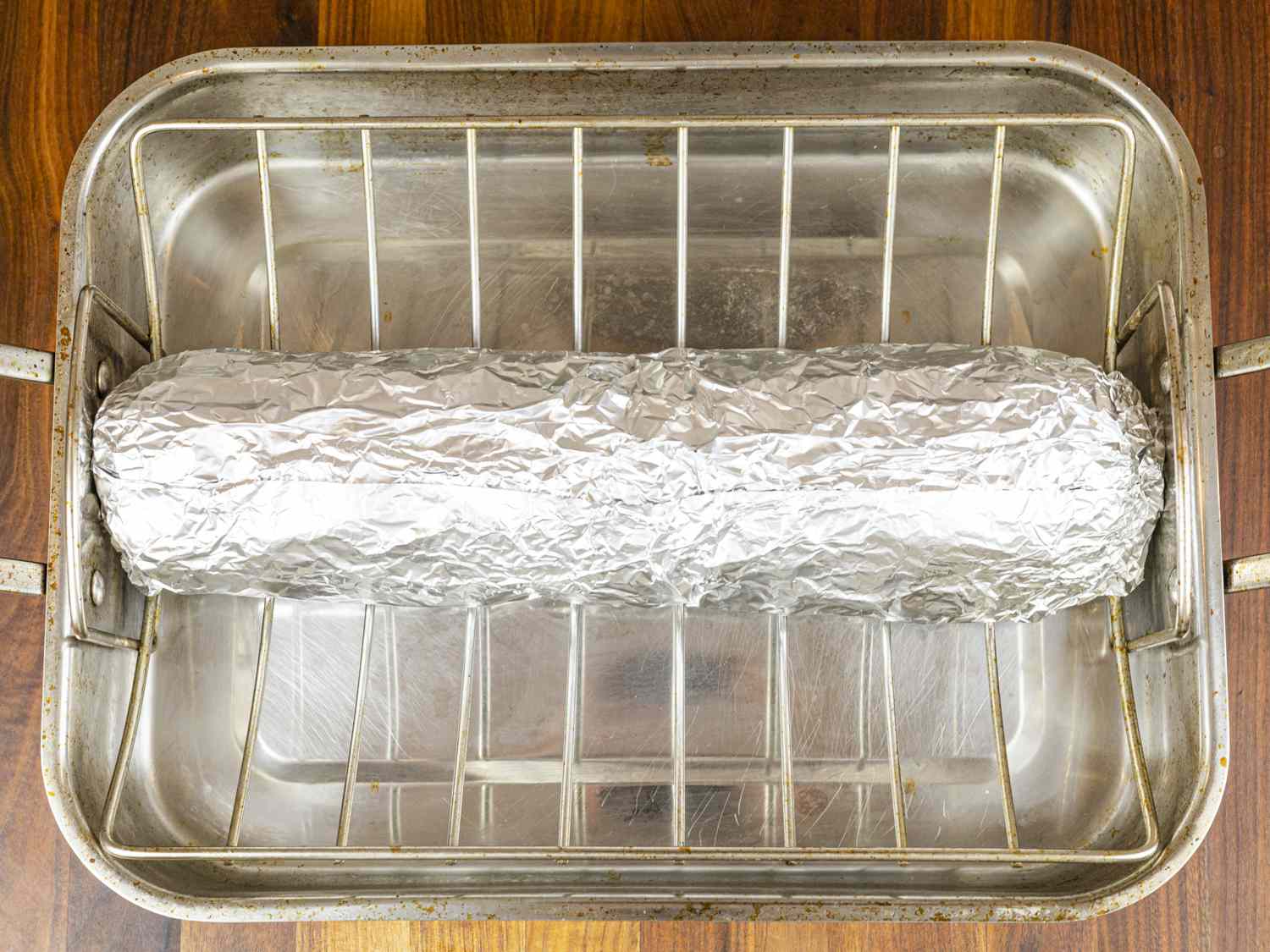 Hands shaping a log of embutido mixture on aluminum foil on a wooden cutting board, with hard-boiled eggs and Vienna sausages lined up for filling