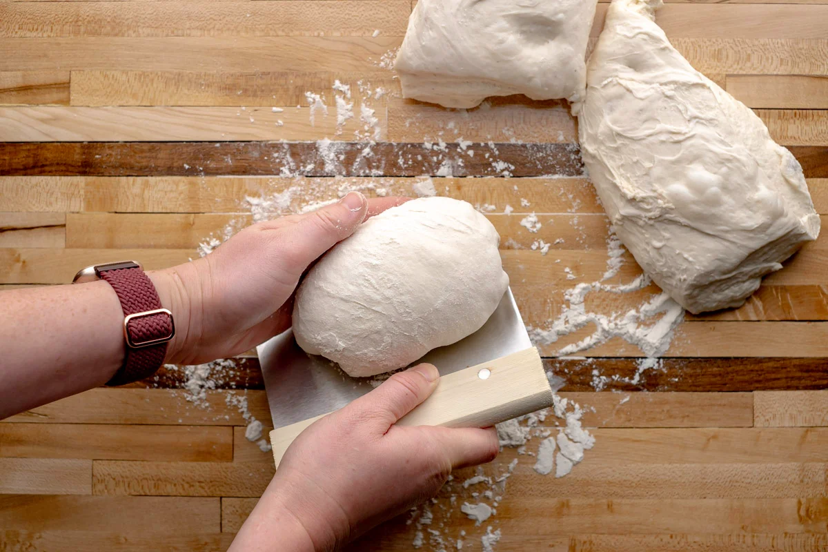 Hands shaping a long baguette loaf on a lightly floured wooden counter with a bench scraper nearby