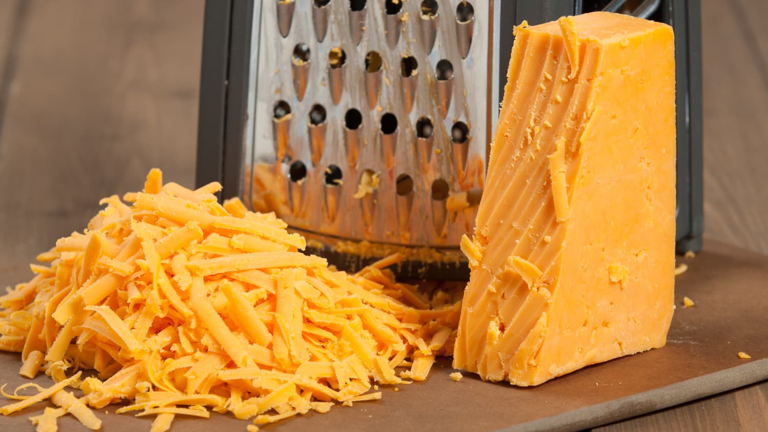 Hands shredding a block of sharp cheddar on a box grater over a cutting board in a home kitchen