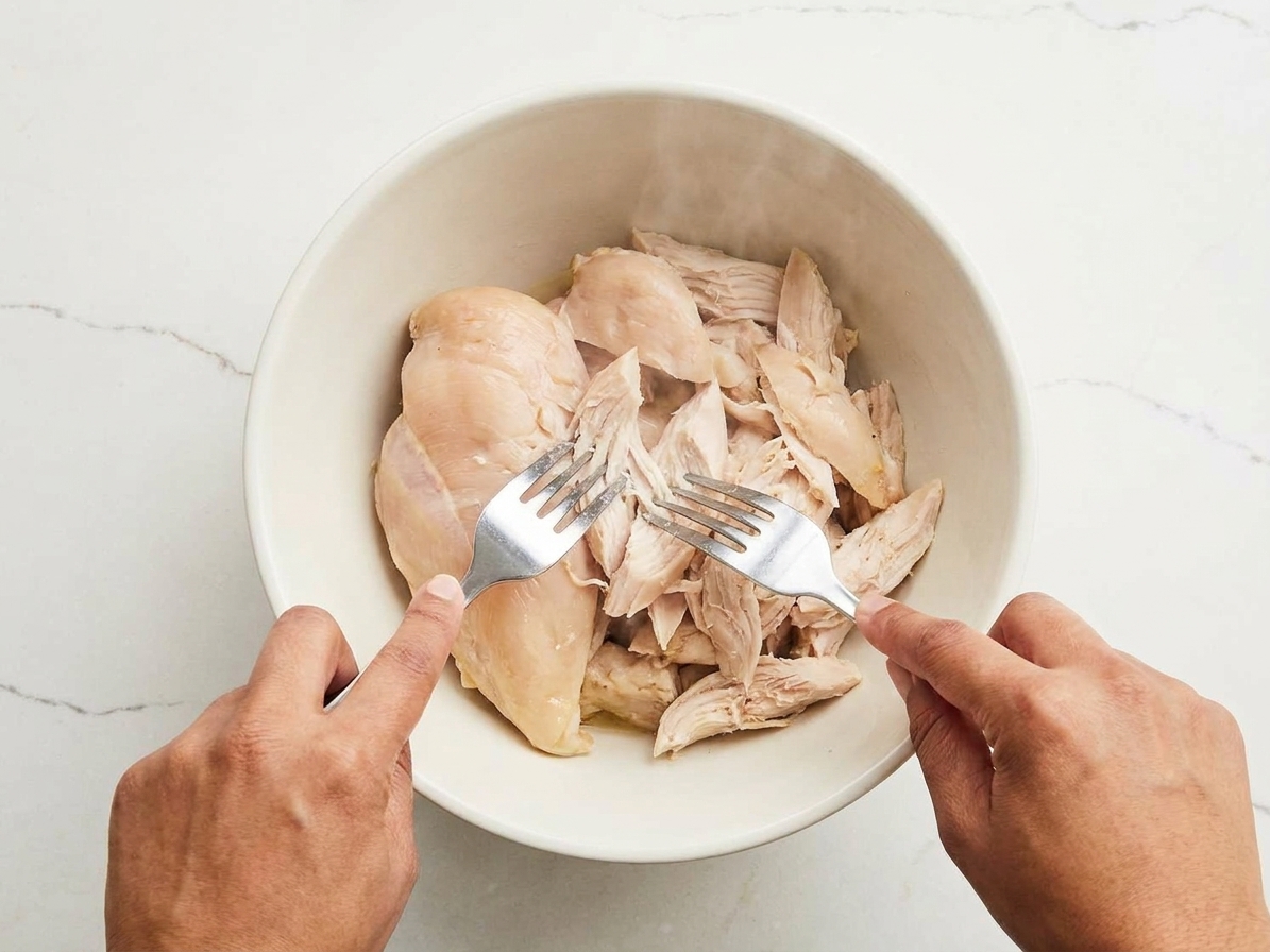 Hands shredding cooked chicken with two forks in a bowl on a kitchen counter