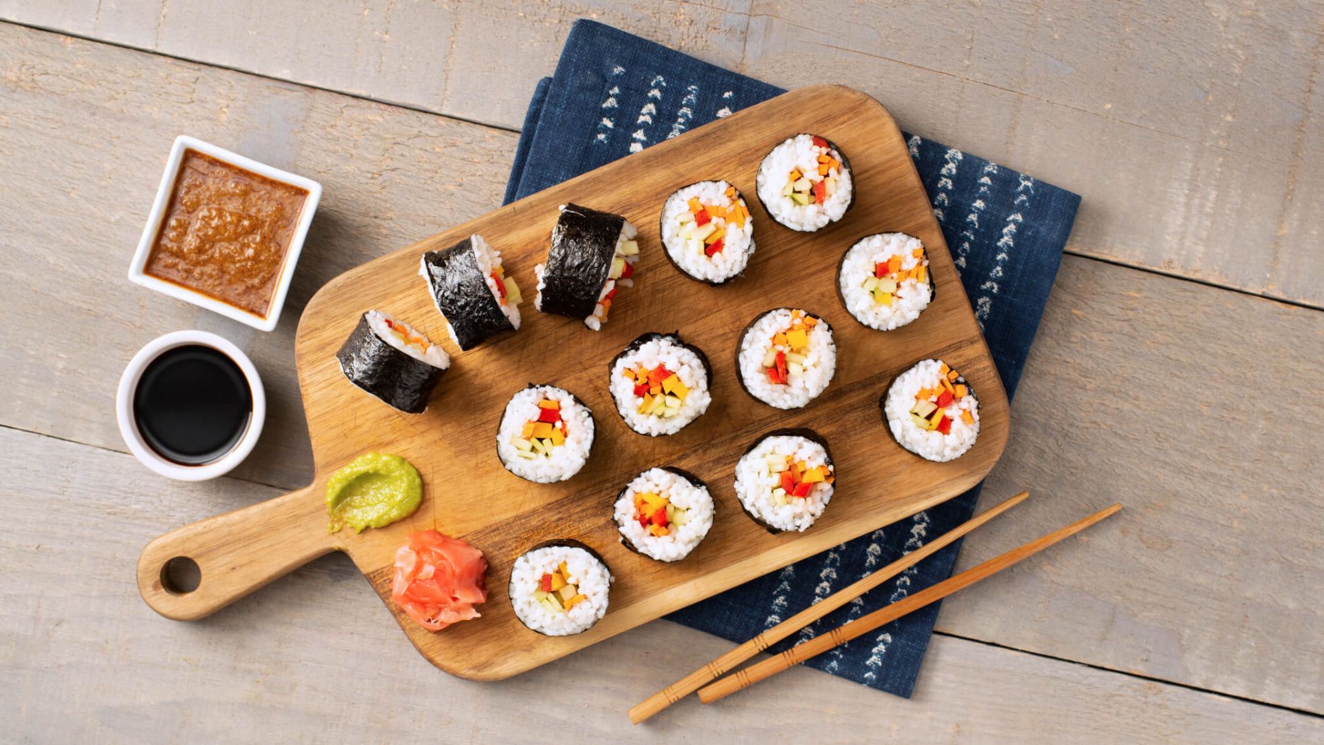 Hands slicing a sushi roll with a sharp chef's knife on a wooden cutting board, with a damp towel nearby for wiping the blade