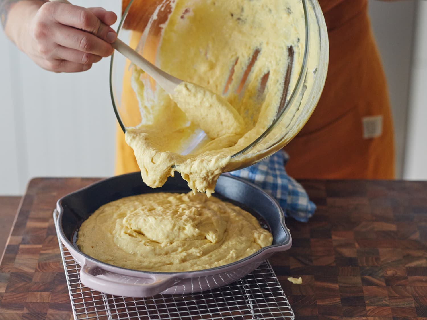 Hands spooning cornbread batter over a saucy meat and bean filling in a baking dish on a kitchen counter