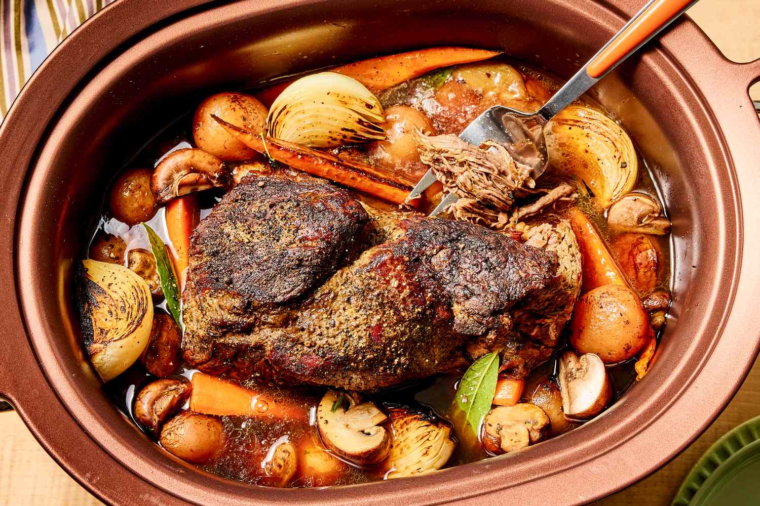 Hands sprinkling salt and black pepper over a beef chuck roast on a cutting board beside sliced onions and carrots
