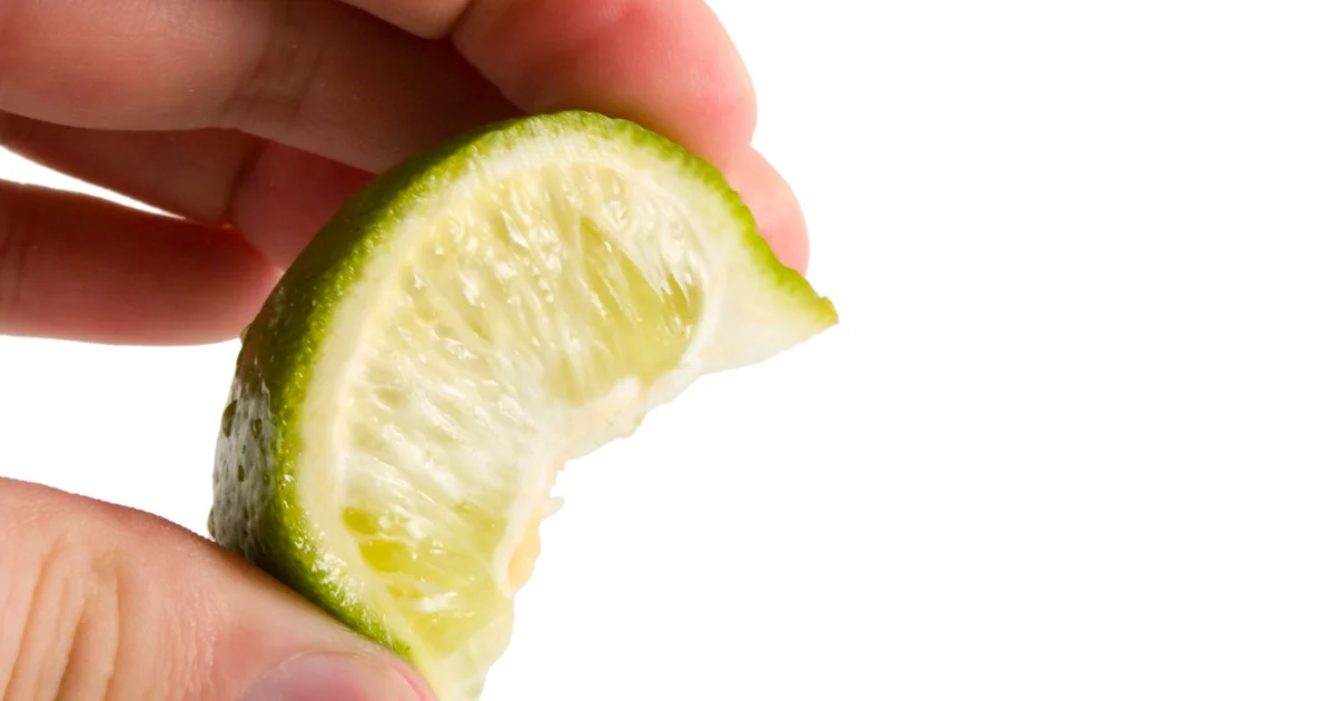Hands squeezing a fresh lime wedge over a glass filled with ice on a kitchen counter