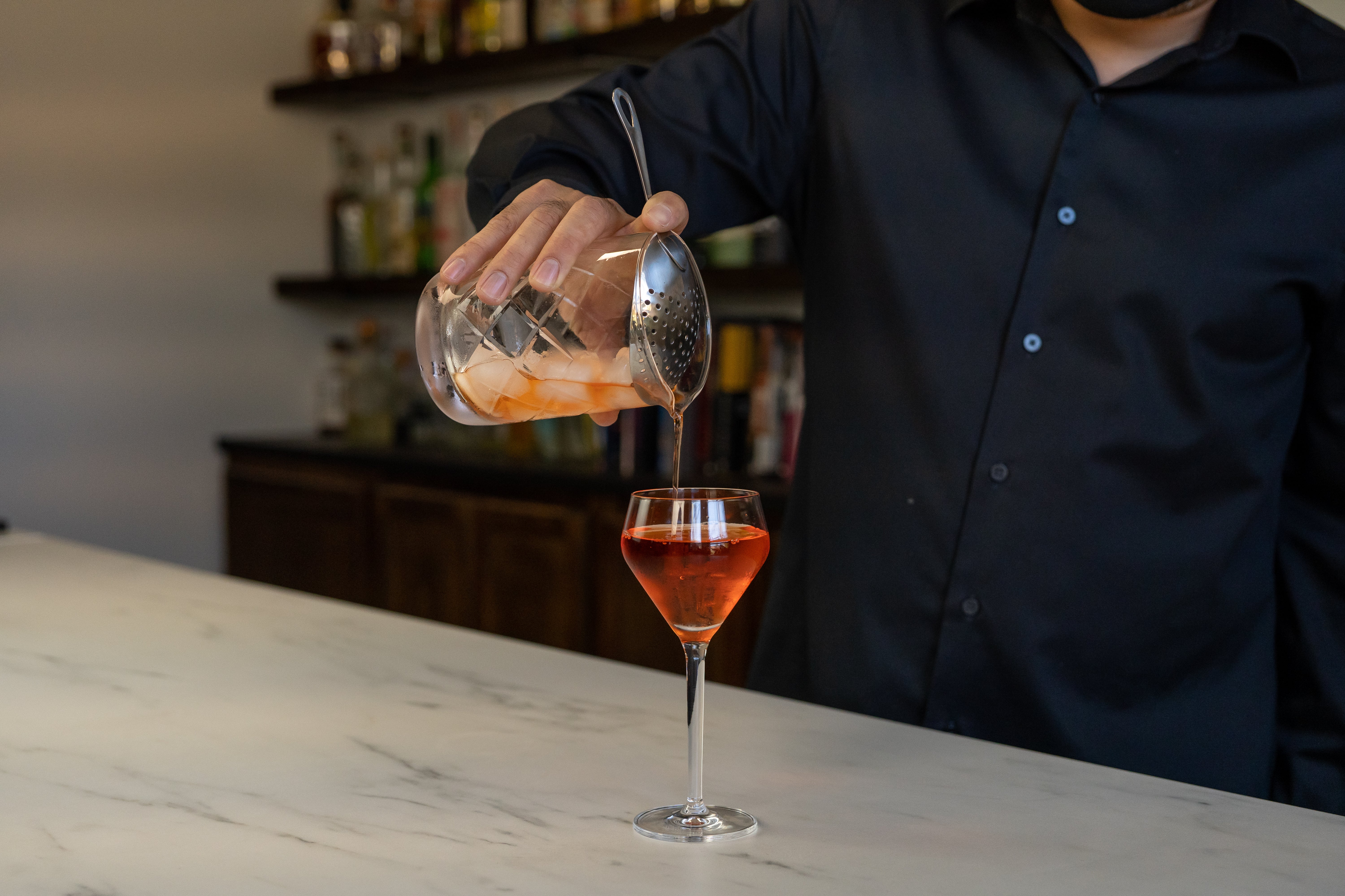 Hands stirring a Manhattan in a mixing glass packed with ice using a long bar spoon on a home kitchen counter