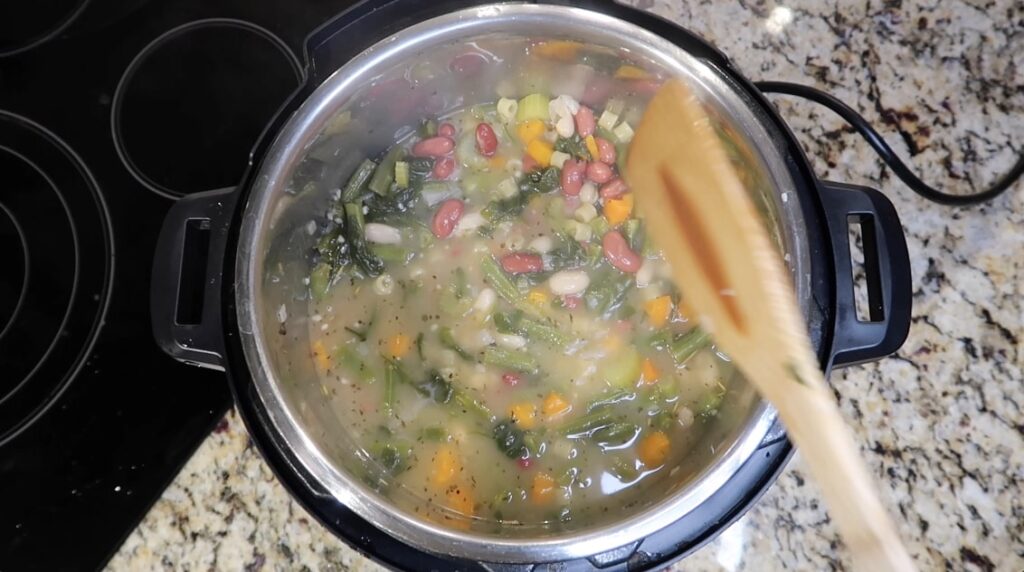 Hands stirring a large pot of simmering minestrone soup on a stovetop with visible vegetables and beans