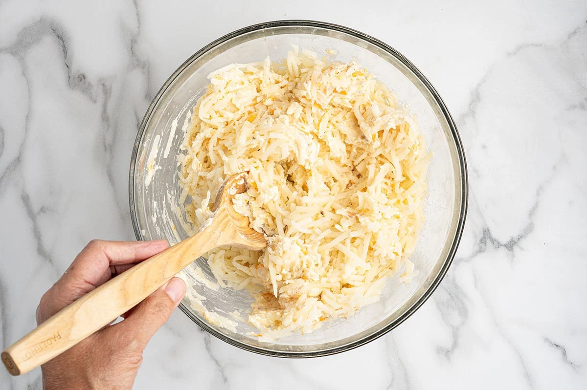 Hands stirring a thick cheesy hashbrown casserole mixture in a large bowl with a rubber spatula on a bright kitchen counter