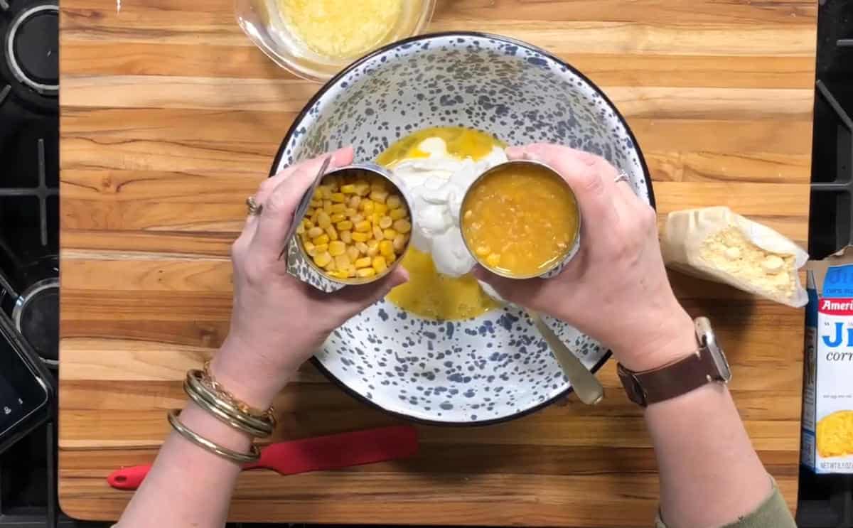 Hands stirring corn pudding batter in a large glass bowl with a wooden spoon, corn kernels visible, on a clean kitchen counter