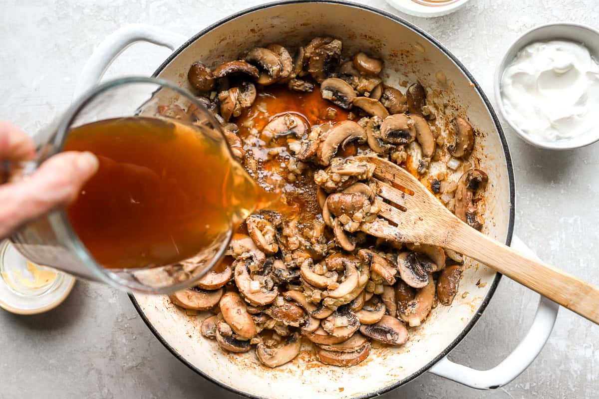 Hands stirring creamy chicken stroganoff in a large skillet on a stovetop, with steam rising