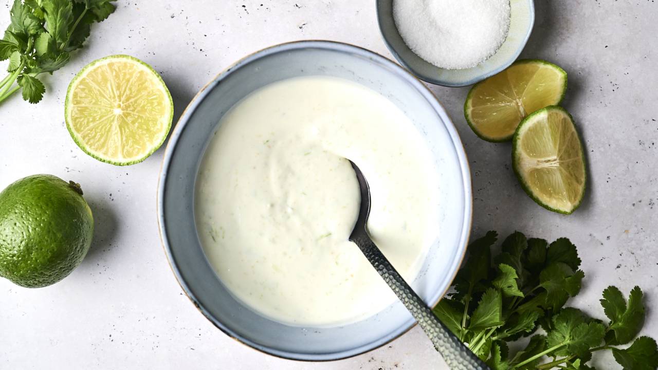 Hands stirring sour cream, lime juice, and garlic in a small ceramic bowl with a spoon on a bright kitchen counter