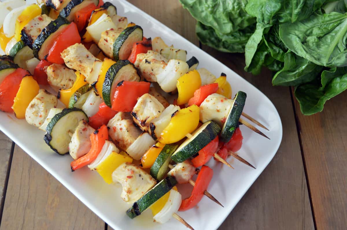 Hands threading marinated chicken, bell pepper, and red onion onto metal skewers over a cutting board on a home kitchen counter