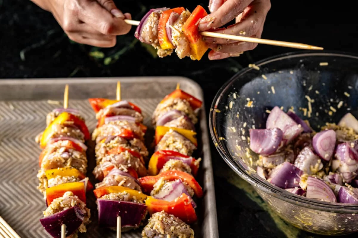Hands threading marinated chicken pieces onto metal skewers on a cutting board with a bowl of marinade nearby