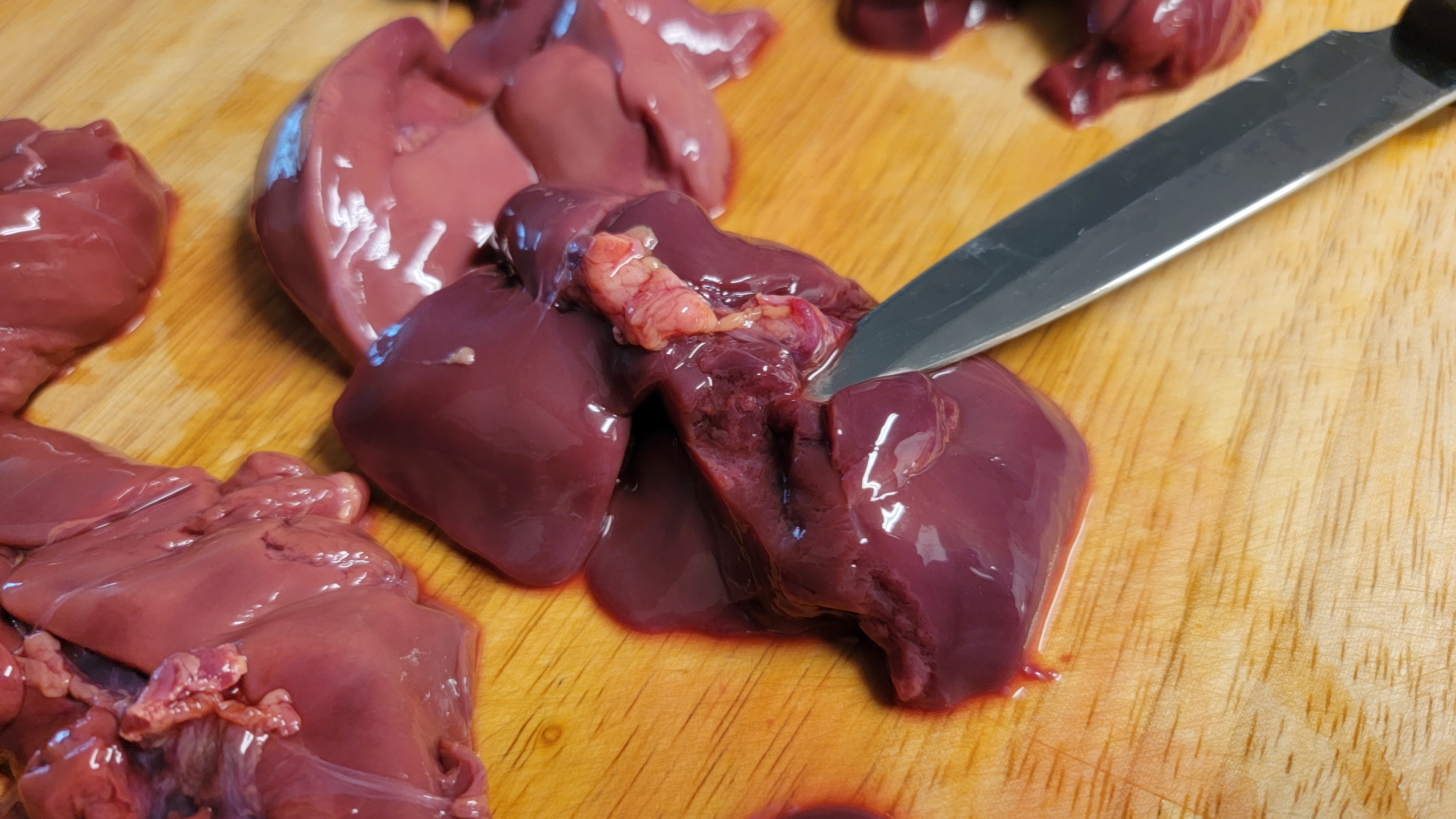 Hands trimming connective tissue from raw chicken livers on a wooden cutting board with a small paring knife