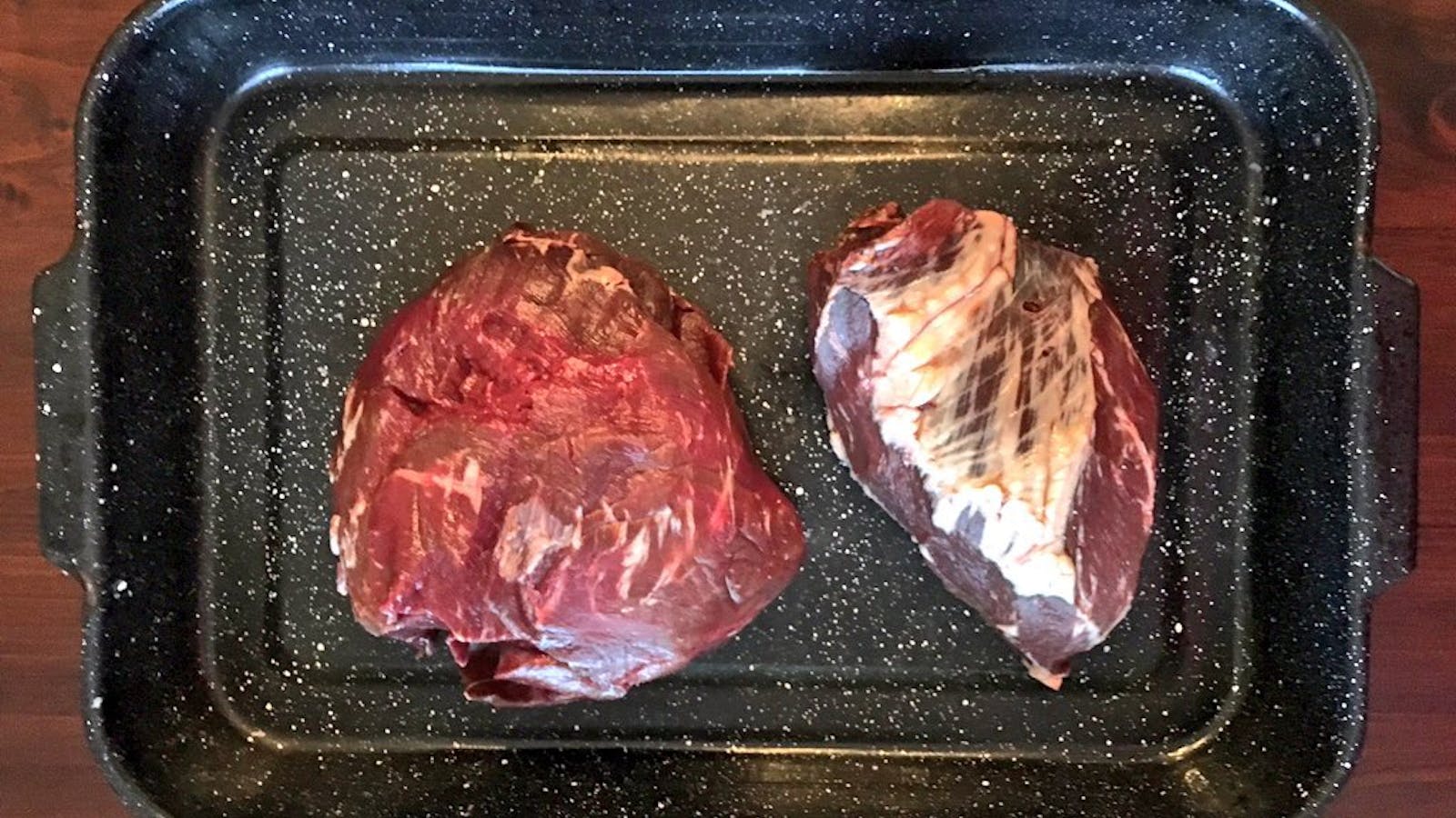 Hands trimming silvery connective tissue from a beef heart with a chef's knife on a wooden cutting board in a home kitchen