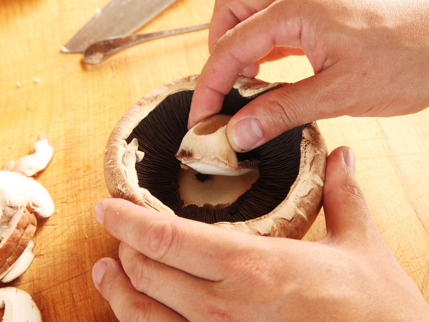 Hands twisting stems out of cremini mushrooms over a cutting board on a bright kitchen counter