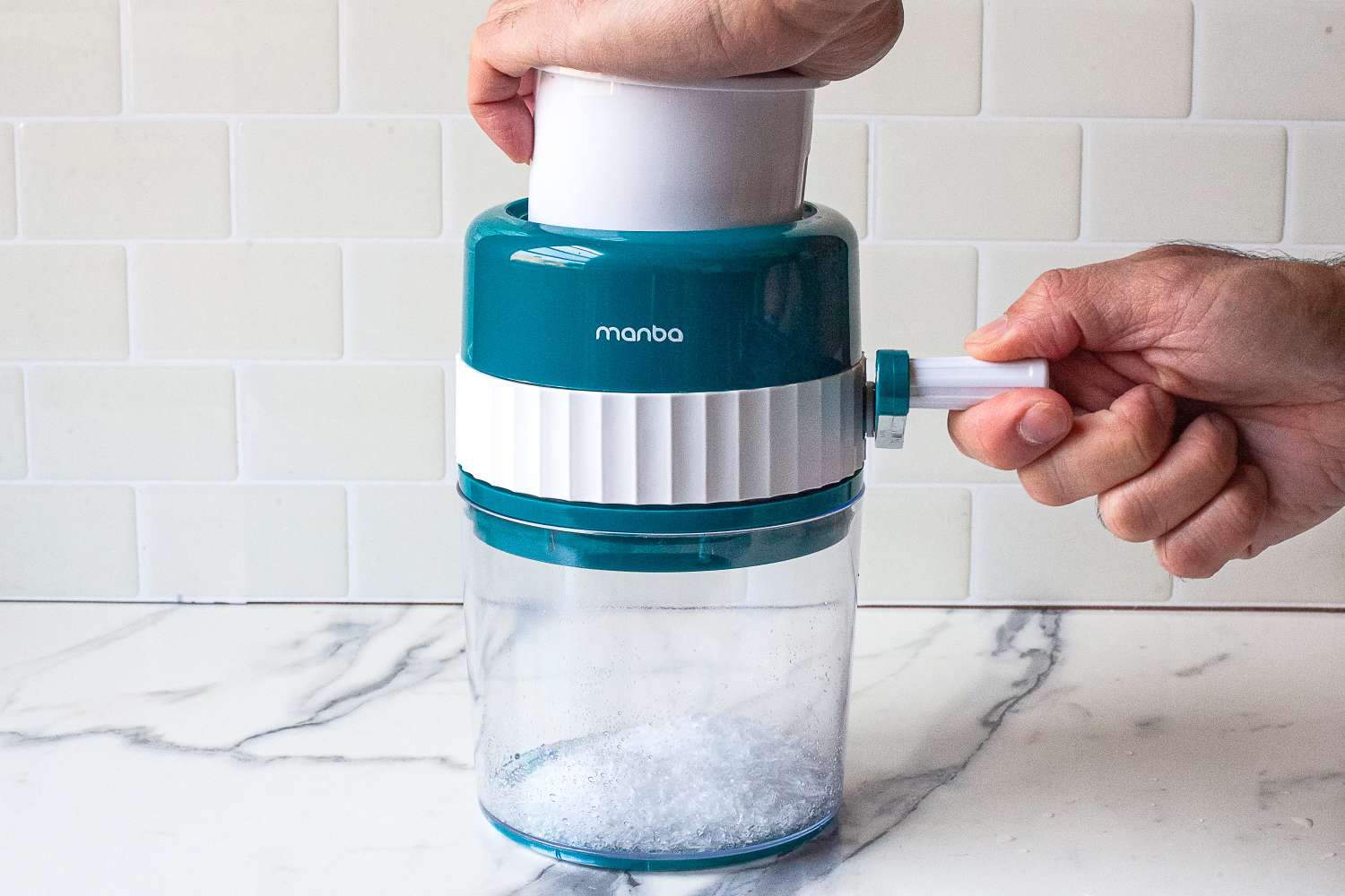 Hands using a countertop ice shaver to make a fluffy pile of shaved ice in a metal bowl