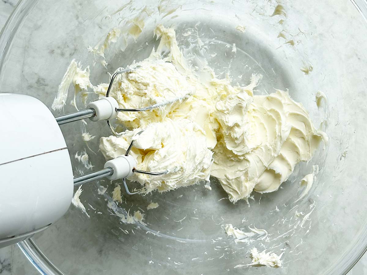 Hands using a hand mixer to beat softened cream cheese in a glass mixing bowl on a kitchen counter with warm natural light