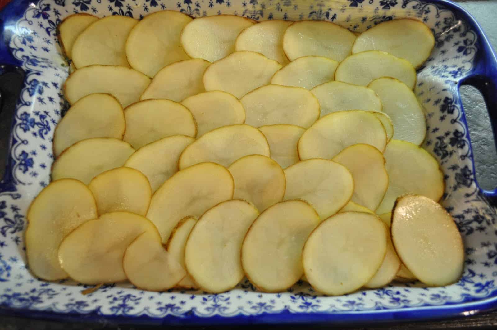Hands using a mandoline to slice peeled potatoes into thin, even rounds on a kitchen counter
