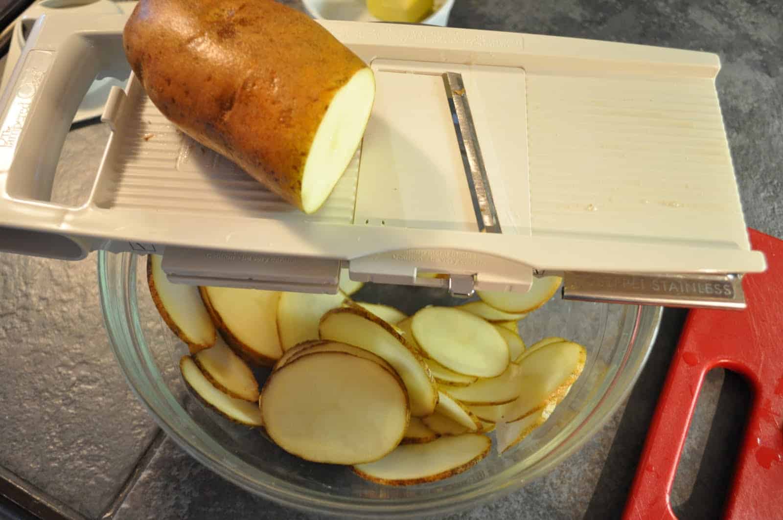 Hands using a mandoline to slice peeled potatoes into thin, even rounds over a bowl on a home kitchen counter