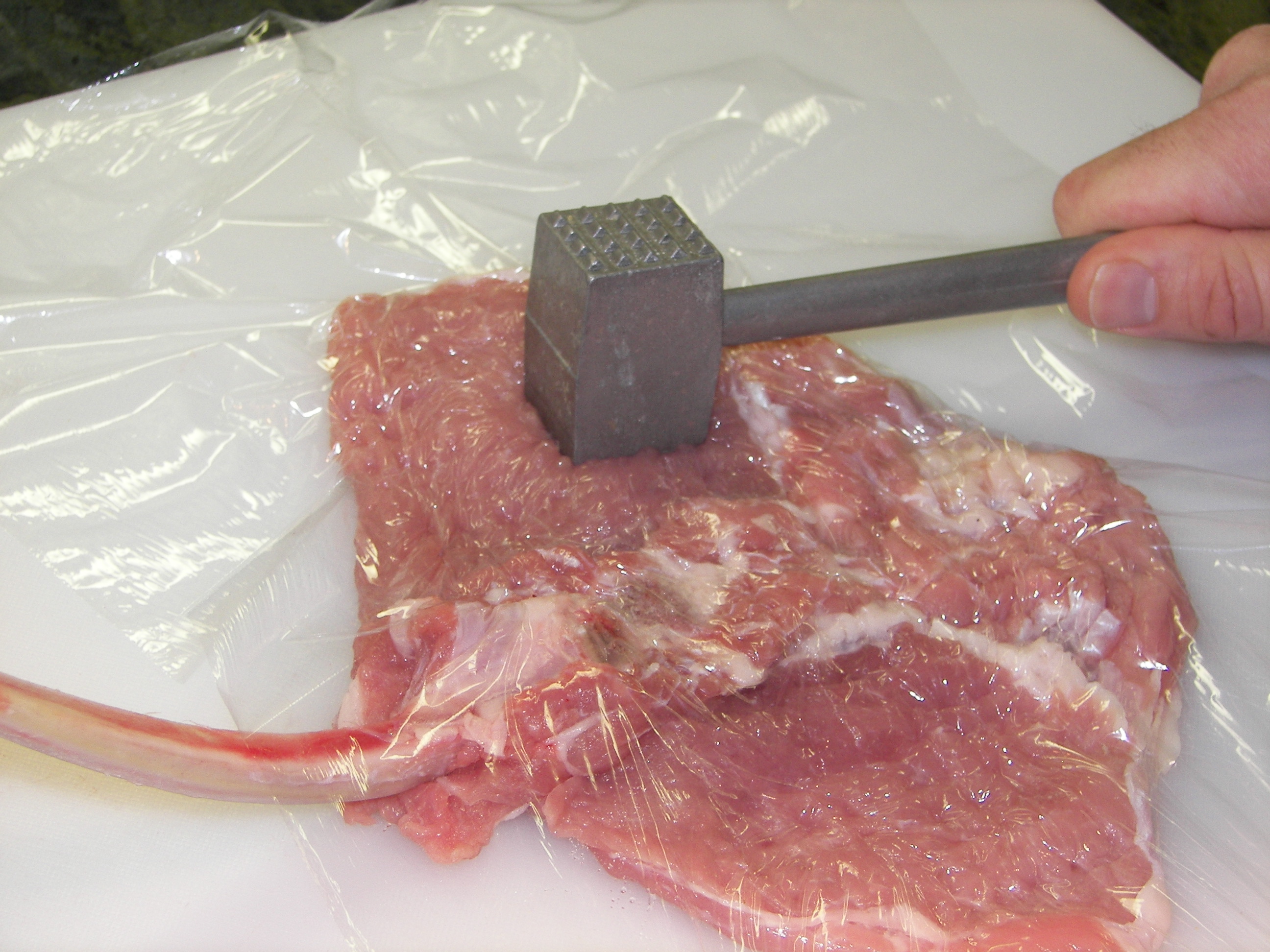 Hands using a meat mallet to pound a veal cutlet thin between parchment paper on a wooden cutting board