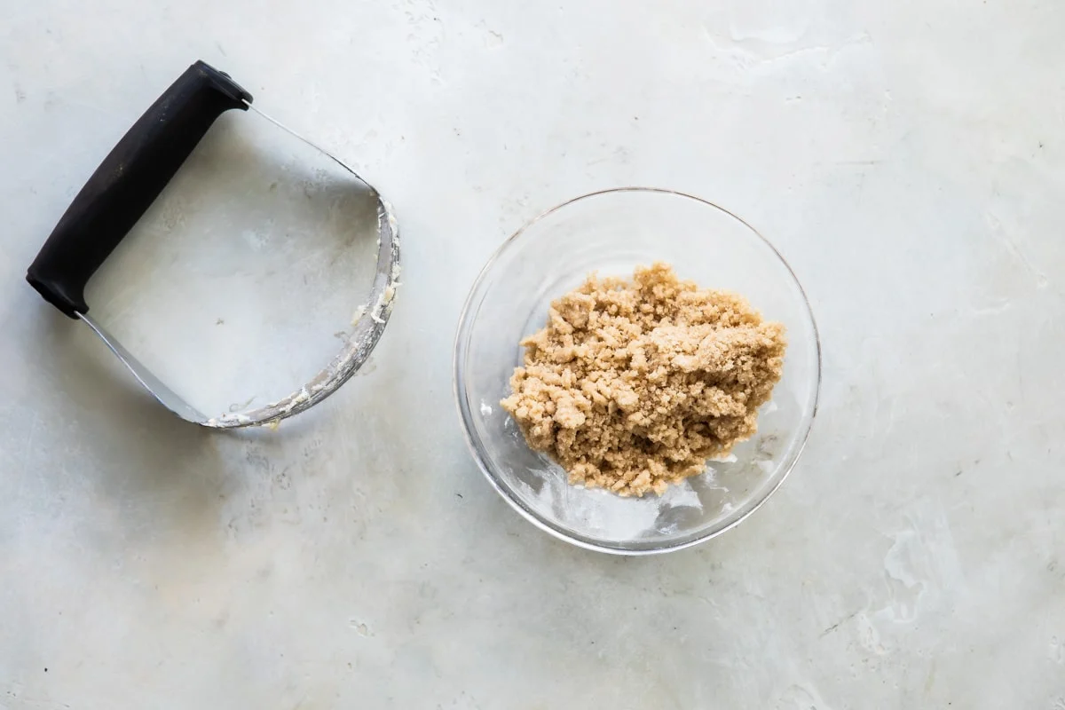 Hands using a pastry cutter in a mixing bowl to cut cold butter into flour and sugar, forming crumbly streusel
