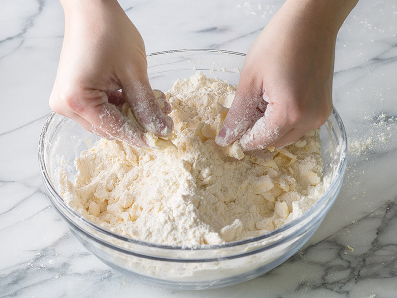 Hands using a pastry cutter to cut cold butter and lard into flour in a large mixing bowl on a kitchen counter
