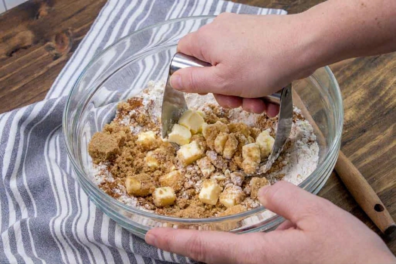 Hands using a pastry cutter to cut cold butter into brown sugar, flour, and cinnamon to form crumbly streusel in a mixing bowl