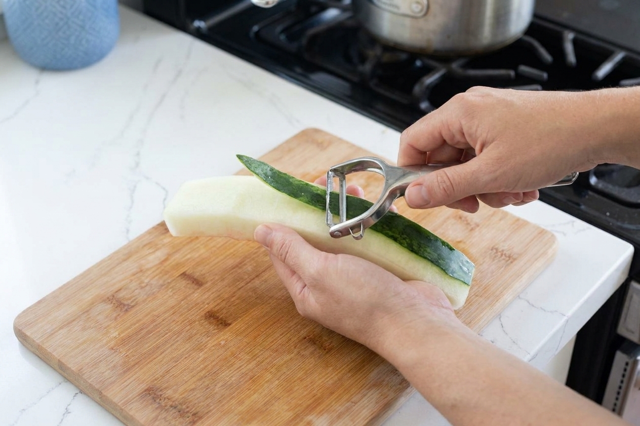 Hands using a vegetable peeler to remove the dark green skin from a strip of watermelon rind on a cutting board