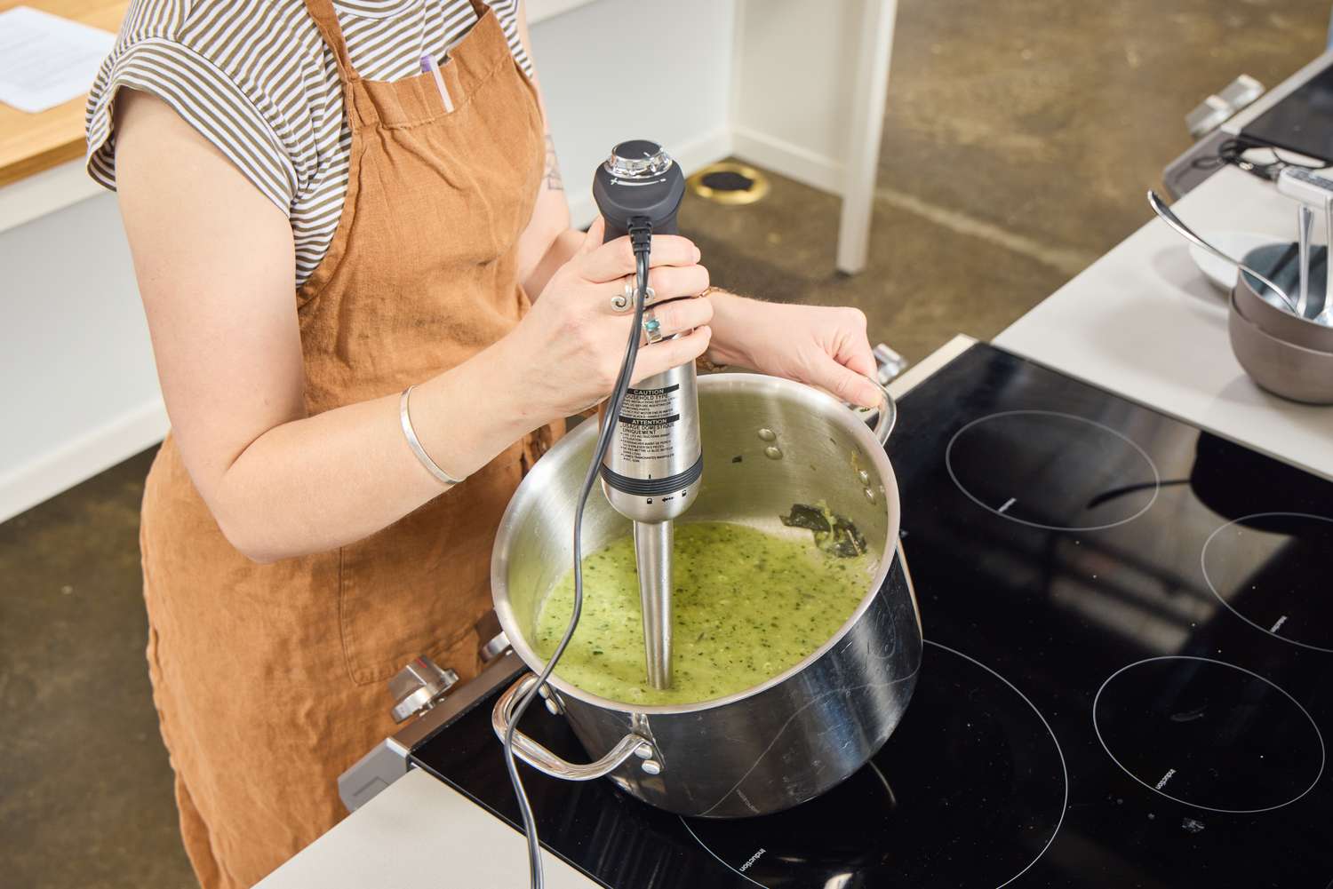 Hands using an immersion blender in a pot of green asparagus soup on a stovetop, steam rising under warm kitchen lighting