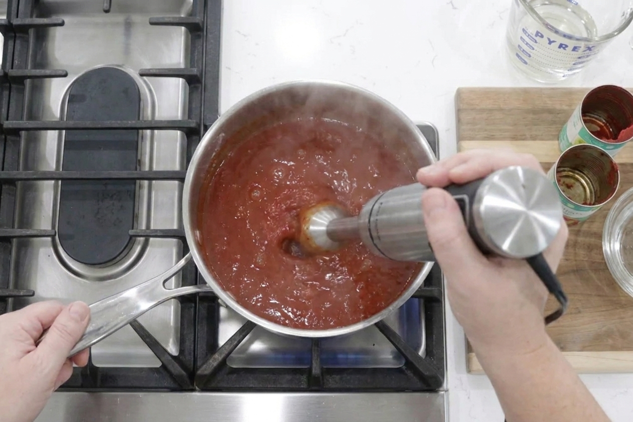 Hands using an immersion blender to puree warm ketchup in a saucepan on a kitchen counter