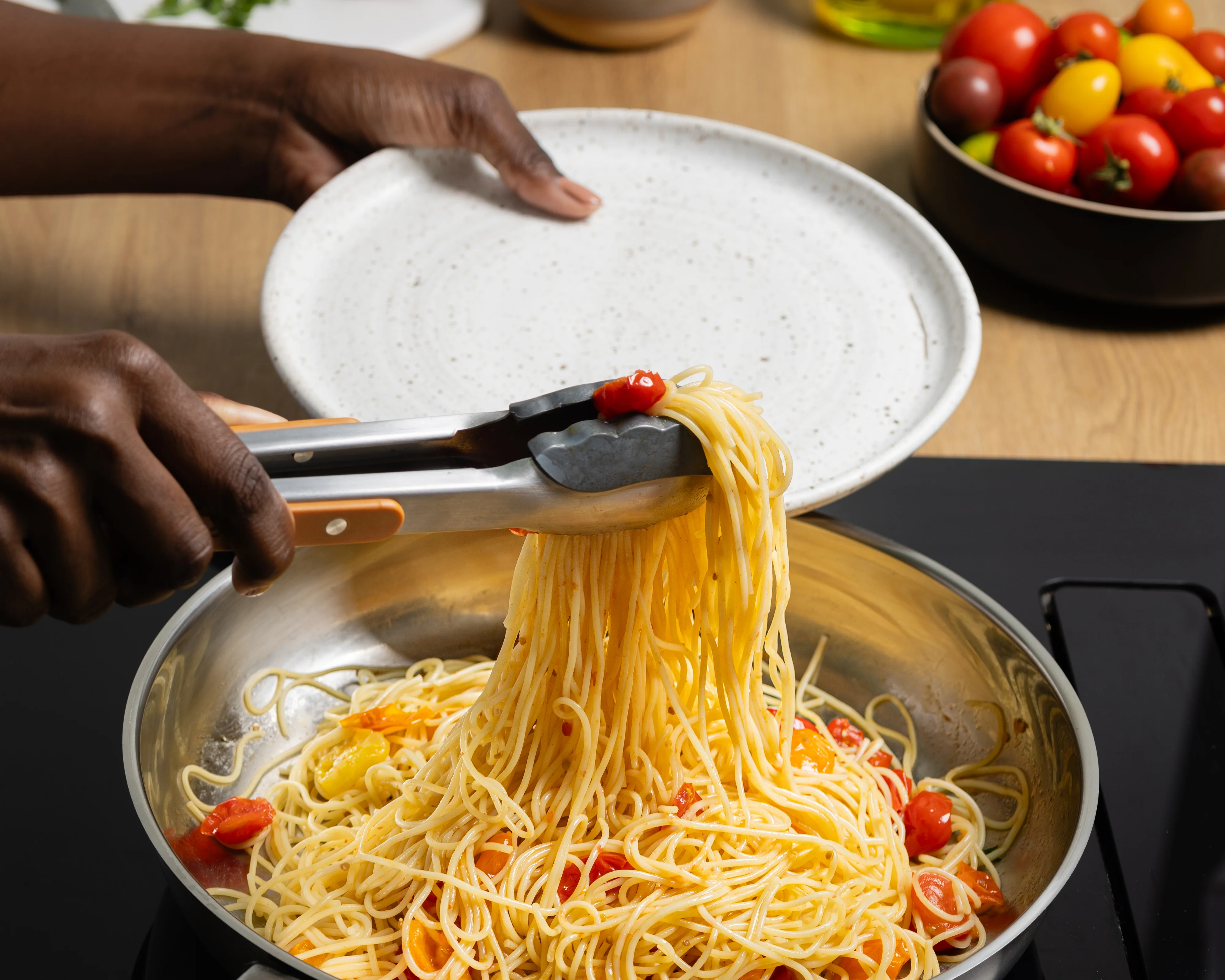 Hands using metal tongs to toss saucy noodles in a large skillet on a home stove