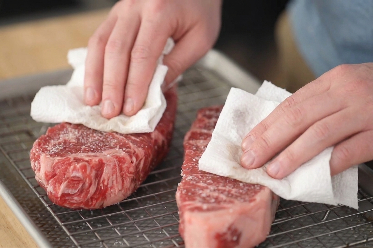 Hands using paper towels to thoroughly pat the surface of a dry-brined steak dry on a wire rack over a baking sheet