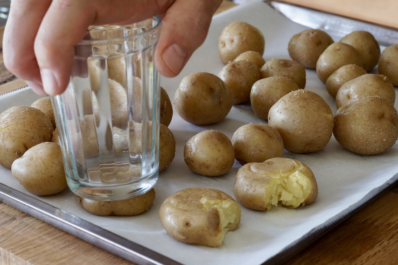 Hands using the bottom of a glass to smash boiled Yukon Gold potatoes on an oiled sheet pan on a kitchen counter