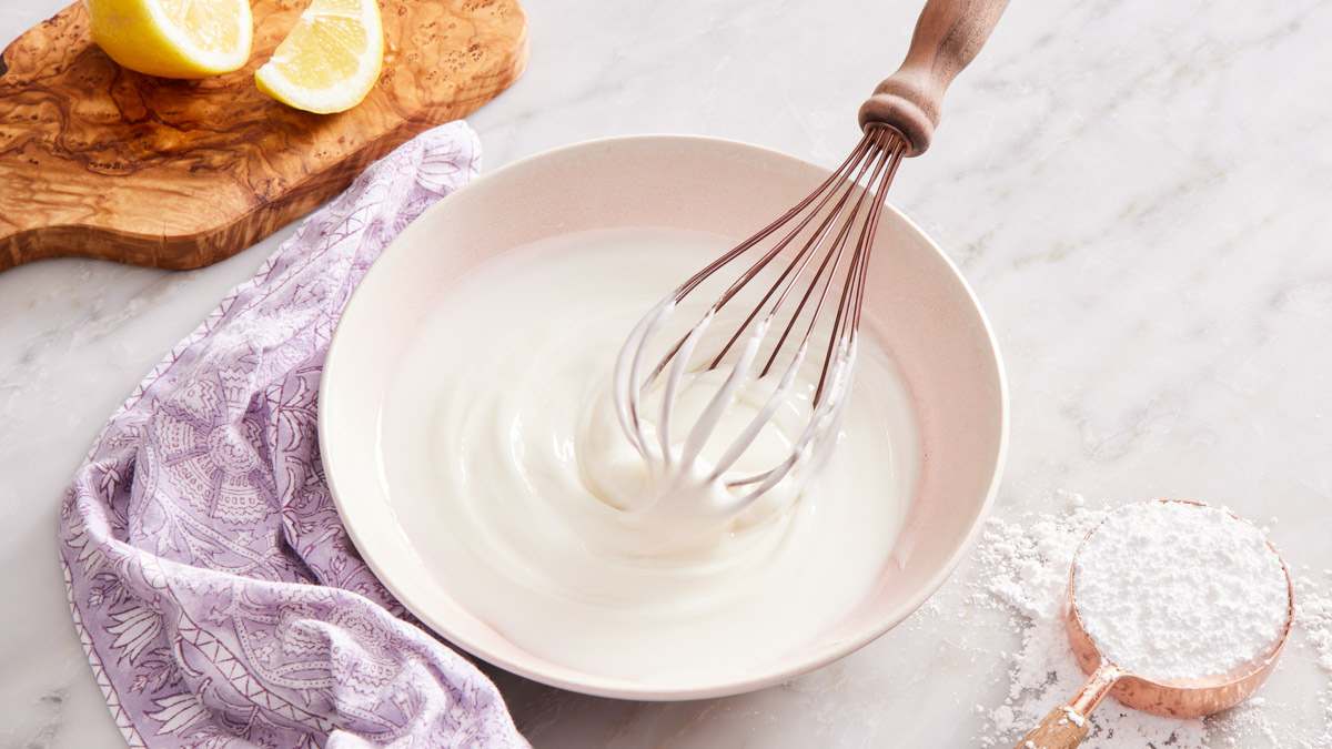 Hands whisking a small bowl of pale yellow lemon icing with a metal whisk on a kitchen counter, with a lemon half nearby