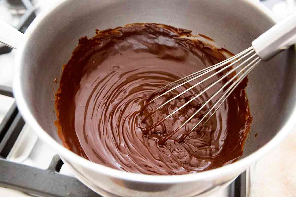 Hands whisking chocolate cake batter in a large glass bowl on a kitchen counter with cocoa powder dusted nearby