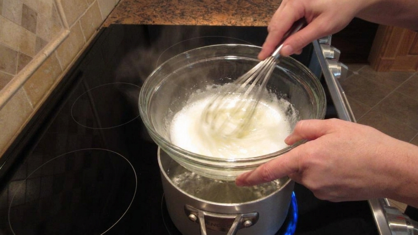 Hands whisking egg whites and sugar in a heatproof bowl over simmering water on a stovetop, with light steam rising