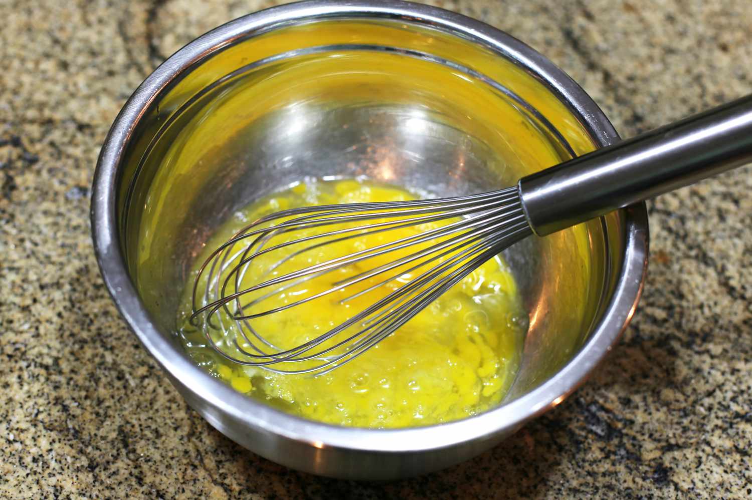 Hands whisking eggs and milk in a large mixing bowl on a kitchen counter next to a whisk and measuring cup