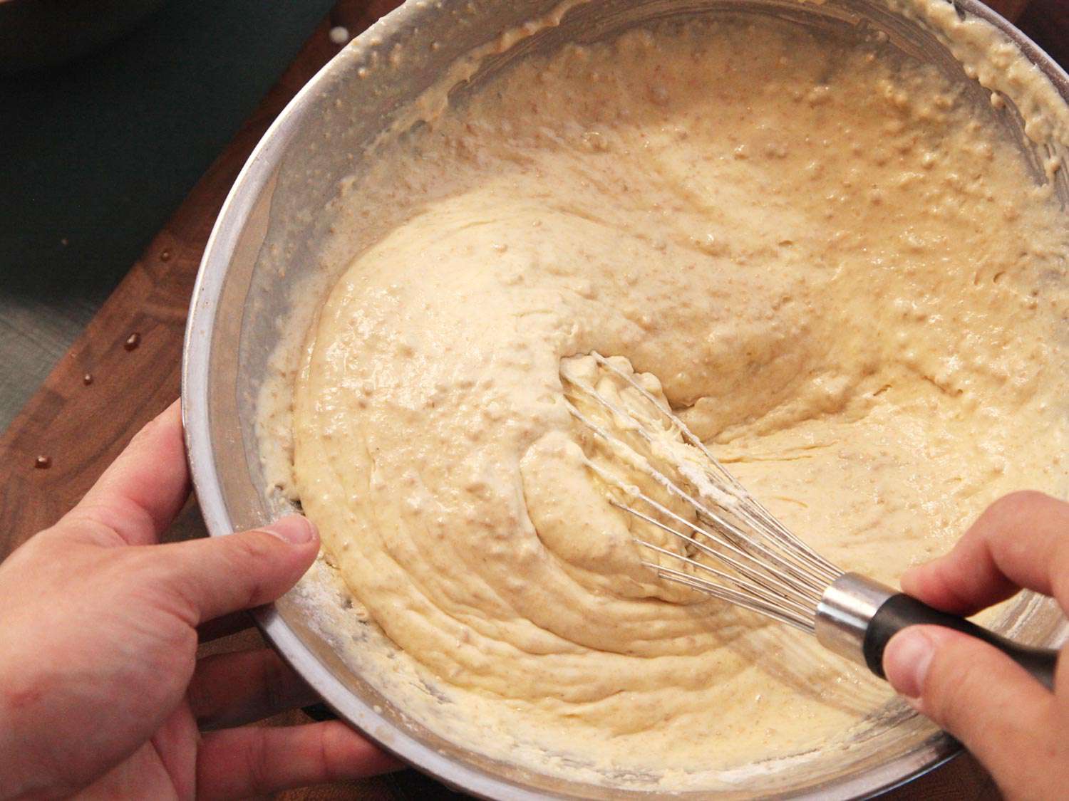 Hands whisking oatmeal pancake batter in a glass bowl on a bright kitchen counter with a measuring cup of milk nearby