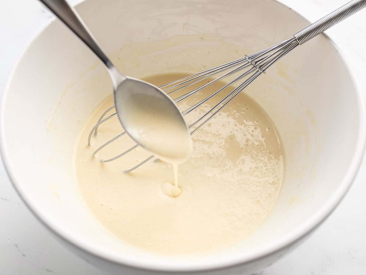 Hands whisking smooth crepe batter in a clear mixing bowl on a light stone kitchen counter with measured ingredients nearby