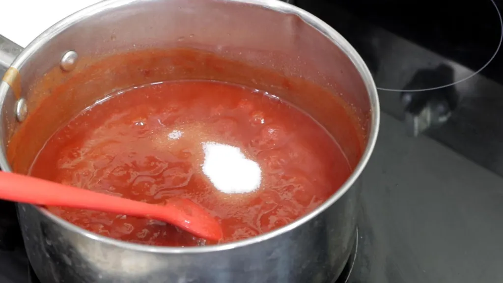 Homemade ketchup gently simmering in a small saucepan with a wooden spoon resting on the rim