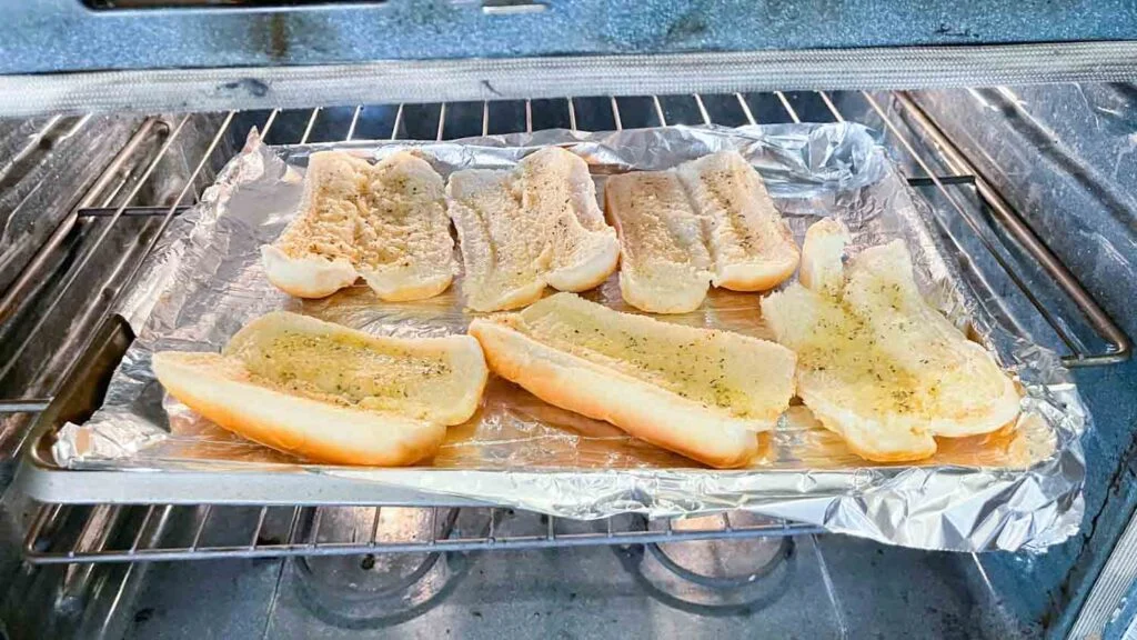 Hot dog buns, butter, minced garlic, parsley, and grated Parmesan arranged on a light kitchen counter