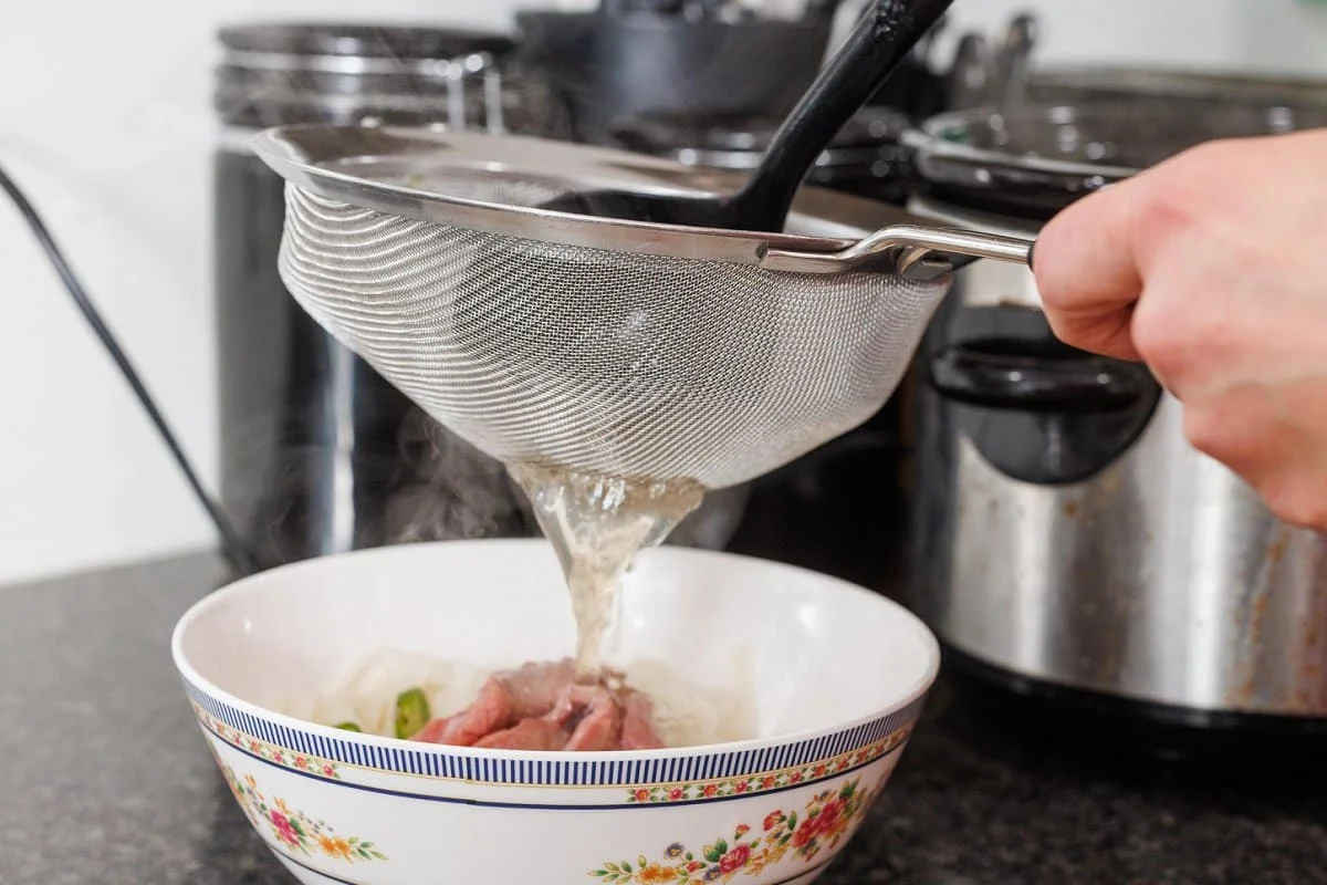 Hot pho broth being poured through a fine mesh strainer into a pot on a stovetop