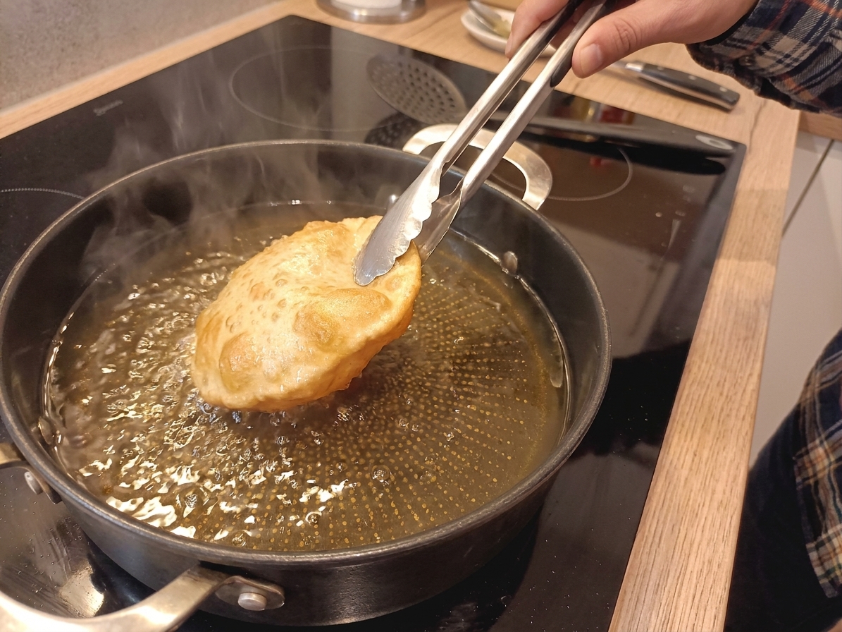 Indian fry bread puffing in hot oil in a cast iron skillet as tongs gently flip it, with golden bubbles forming on the surface