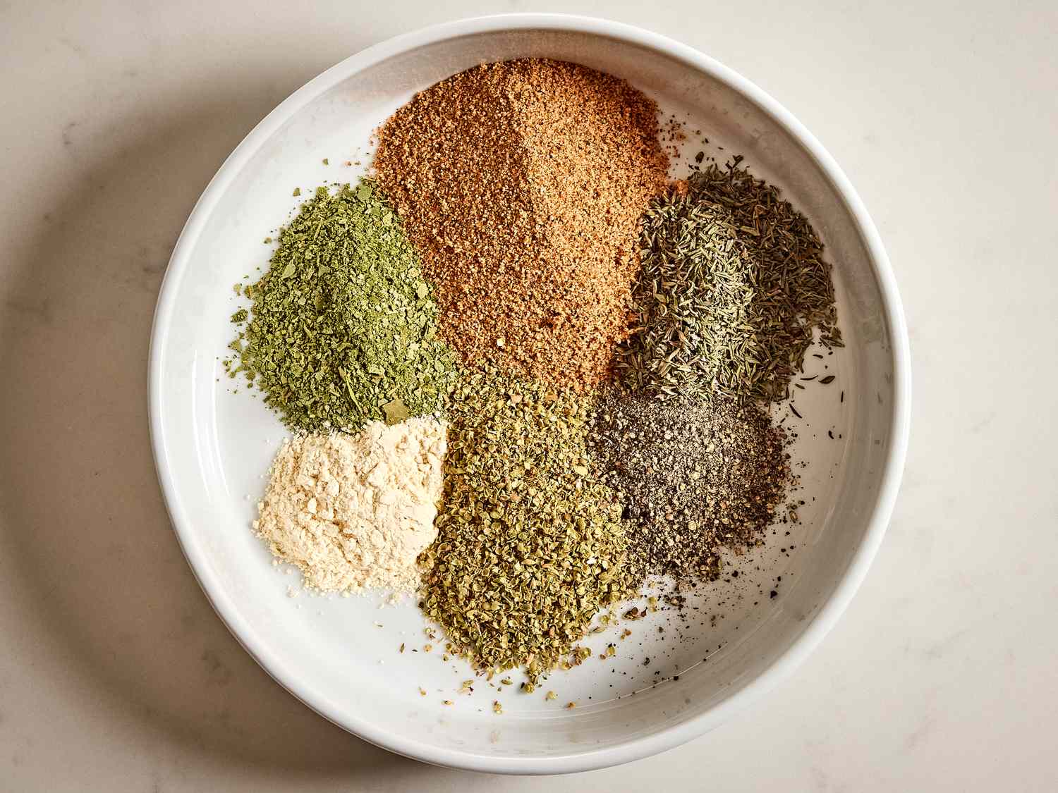 Individual piles of spices for turkey rub on a stone counter, including paprika, brown sugar, salt, pepper, garlic powder, onion powder, and dried herbs in small prep bowls