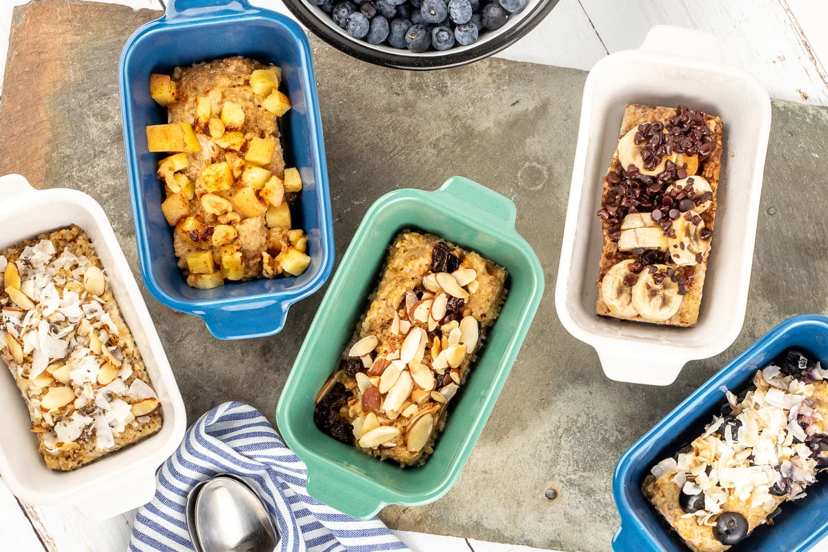 Individual squares of baked oatmeal stacked in a glass meal prep container with parchment paper between layers on a kitchen counter