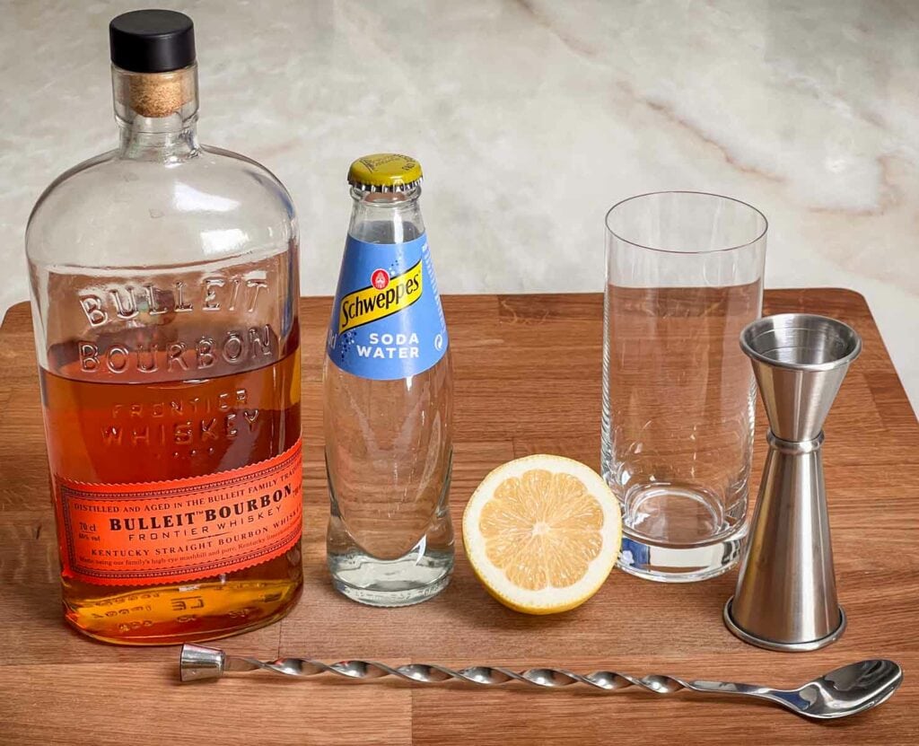 Ingredients for a whiskey highball on a kitchen counter including a bottle of whiskey, a chilled bottle of club soda, a lemon, and a tall highball glass